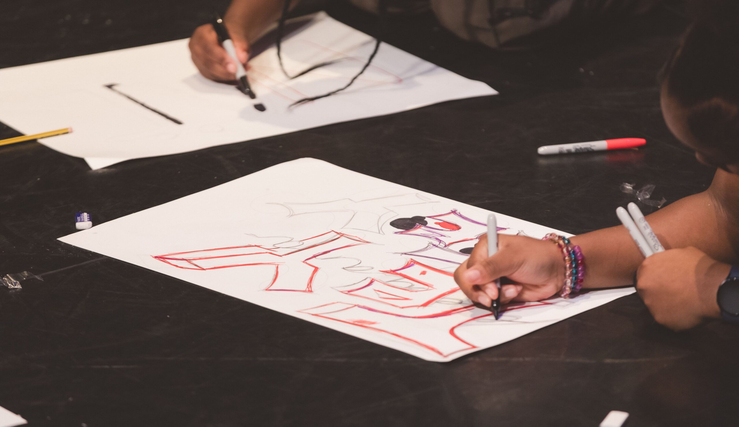 Two young girls graffitiing their name on large white paper with coloured pens.